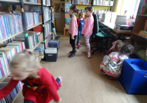Dzieci zwiedzają bibliotekę, oglądają książki na regałach.